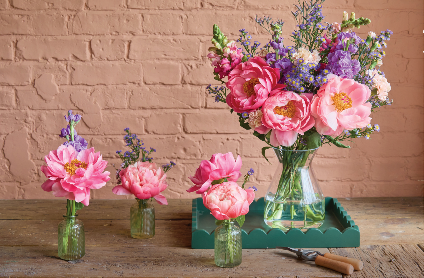 Three small floral arrangements in clear vases on a wooden surface with a brick wall background.